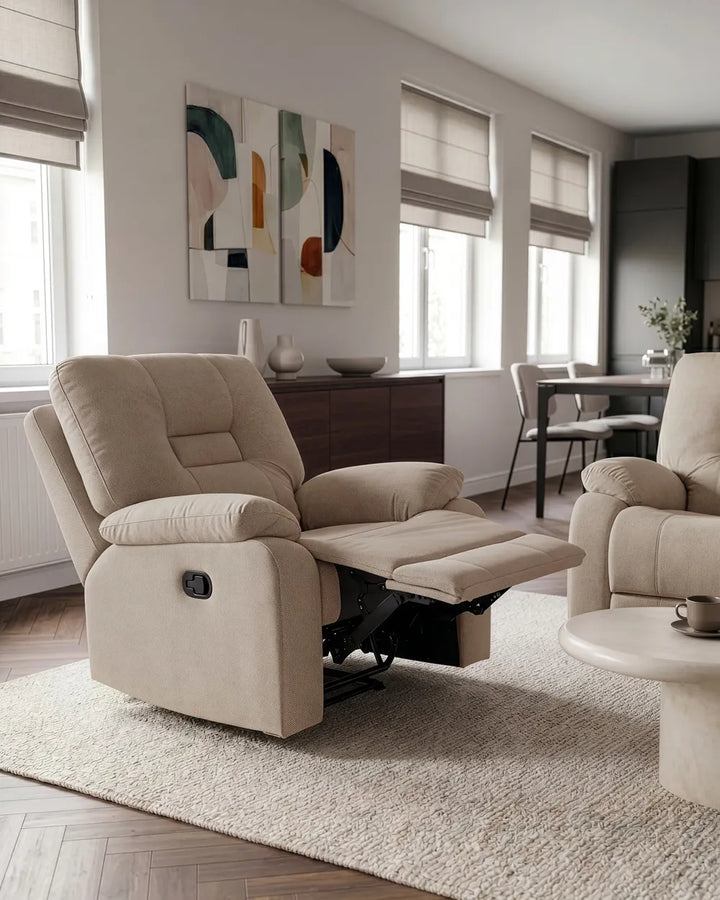 A beige upholstered recliner chair with the footrest extended, shown in a modern living room setting with a matching armchair, rug, windows, and wall art.
