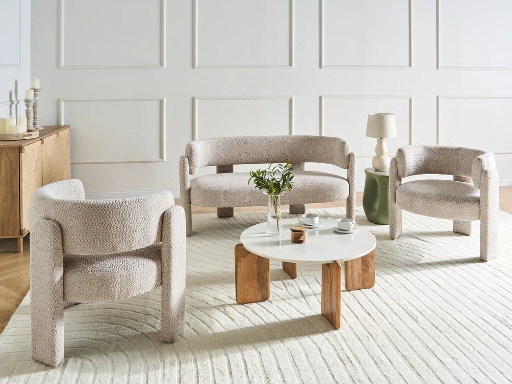 A styled interior scene showing a coordinated furniture set: a curved upholstered loveseat, two matching armchairs, and a round coffee table, arranged on a light rug in a bright room with paneled walls.