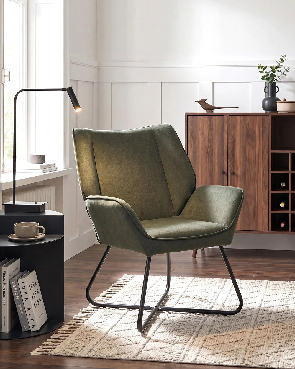 An olive green upholstered accent chair with a black metal sled base, styled in a modern living room setting on a woven rug next to a black side table and lamp.