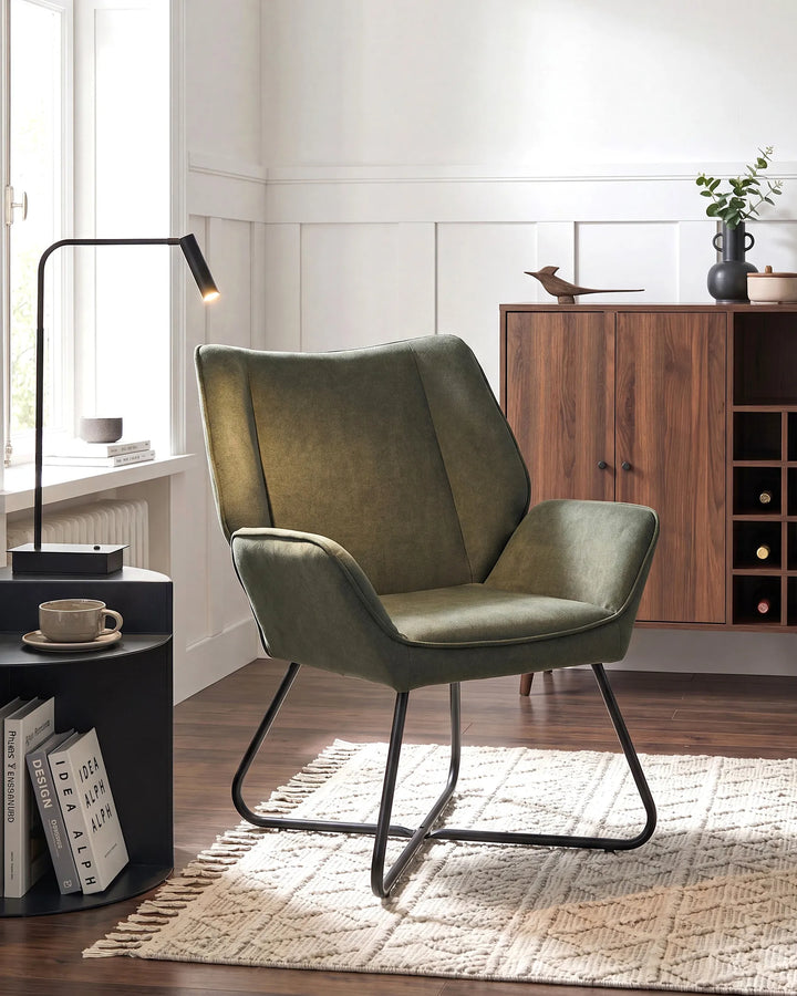 An olive green upholstered accent chair with a black metal sled base, styled in a modern living room setting on a woven rug next to a black side table and lamp.