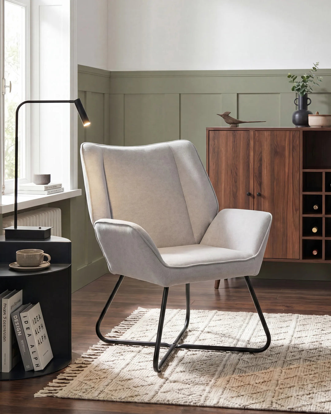 A light beige upholstered accent chair with a black metal frame, styled in a modern living room setting next to a window and a wooden cabinet.