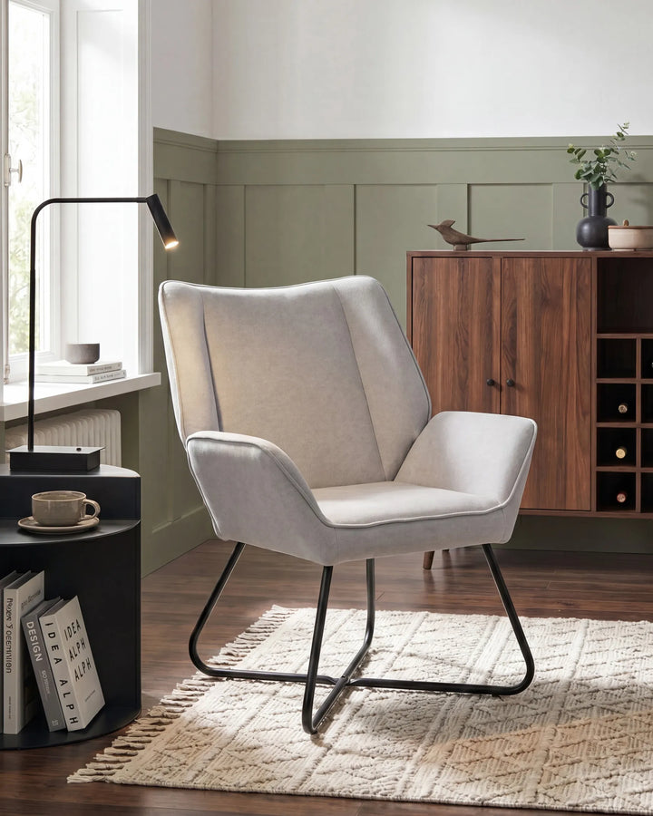 A light beige upholstered accent chair with a black metal frame, styled in a modern living room setting next to a window and a wooden cabinet.