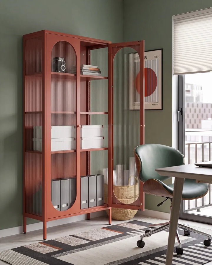 A tall rust-red metal storage cabinet with glass doors and arched upper panels, shown open in a styled home office setting with a green chair, desk, rug, and wall art.