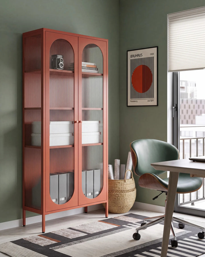 A tall rust-orange metal storage cabinet with two arched glass doors stands in a styled room corner next to a desk and chair.