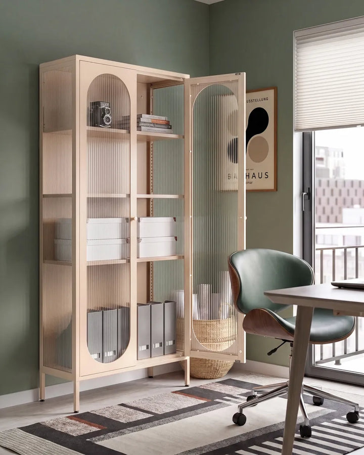 A tall light wood storage cabinet with open shelving and ribbed glass doors stands in a styled home office. The cabinet is shown open with books, boxes, and binders inside, next to a green office chair and desk in a room with sage green walls.