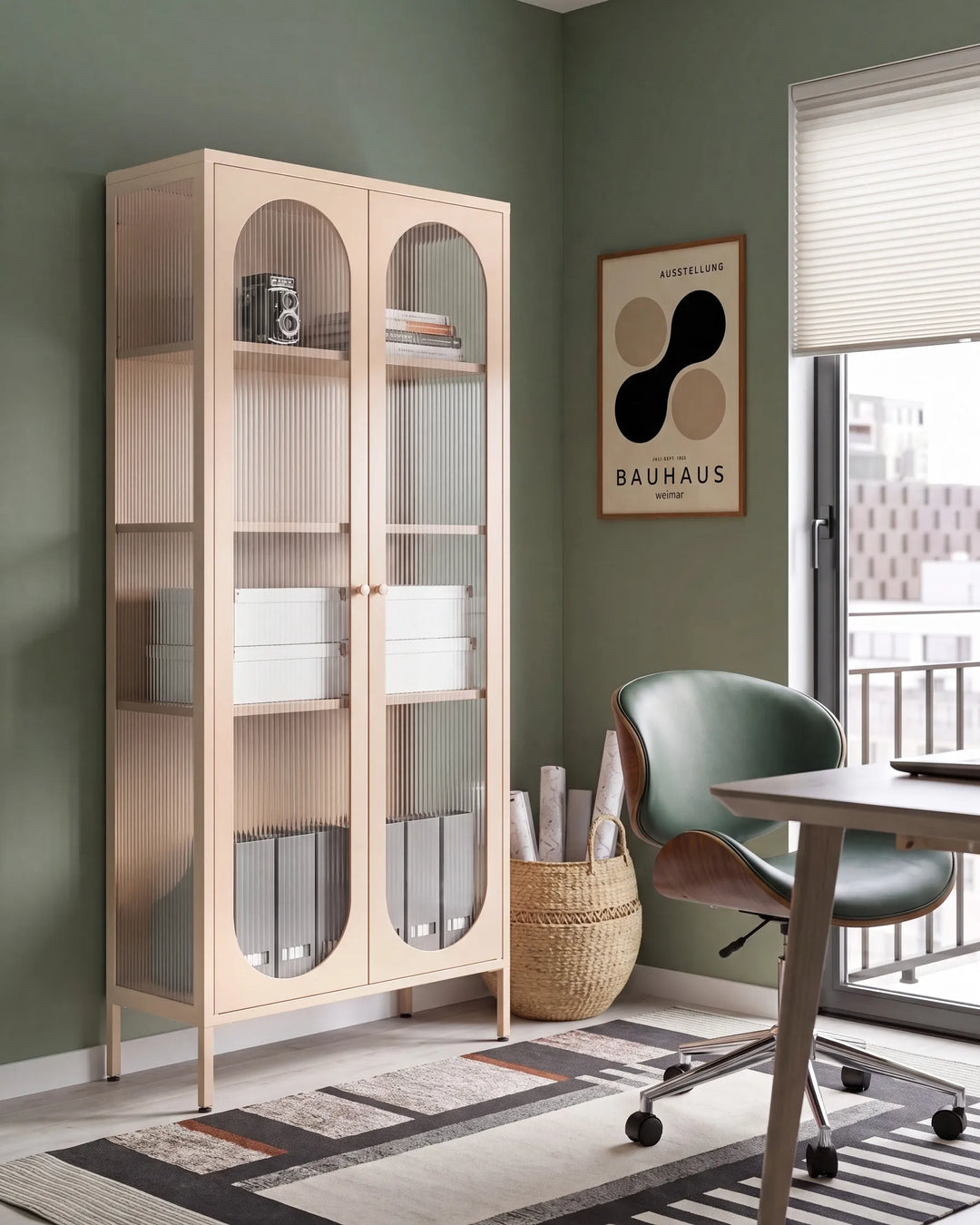 A tall, light beige wooden storage cabinet with two arched glass doors stands in a styled room corner next to a desk and chair. The cabinet features multiple open shelves and ribbed glass panels.