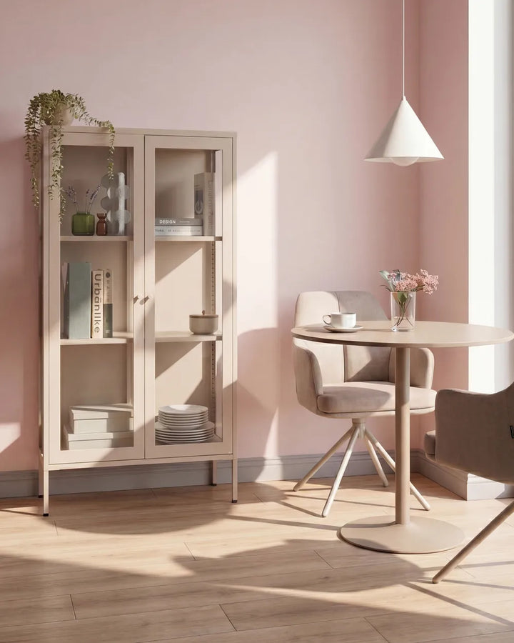A light beige wooden display cabinet with glass doors stands against a dusty pink wall in a sunlit room. The cabinet is styled with books, vases, and a trailing plant.