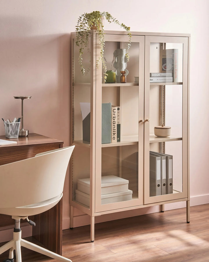 A tall, light beige wooden bookcase with glass doors and shelves, styled in a home office setting next to a desk and chair.
