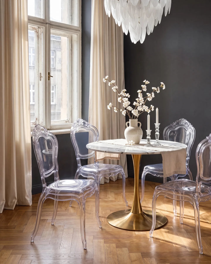 A dining room scene with a round white marble-top table on a gold pedestal base, surrounded by clear acrylic chairs. A white vase with white blossoms and two candlesticks sit on the table.