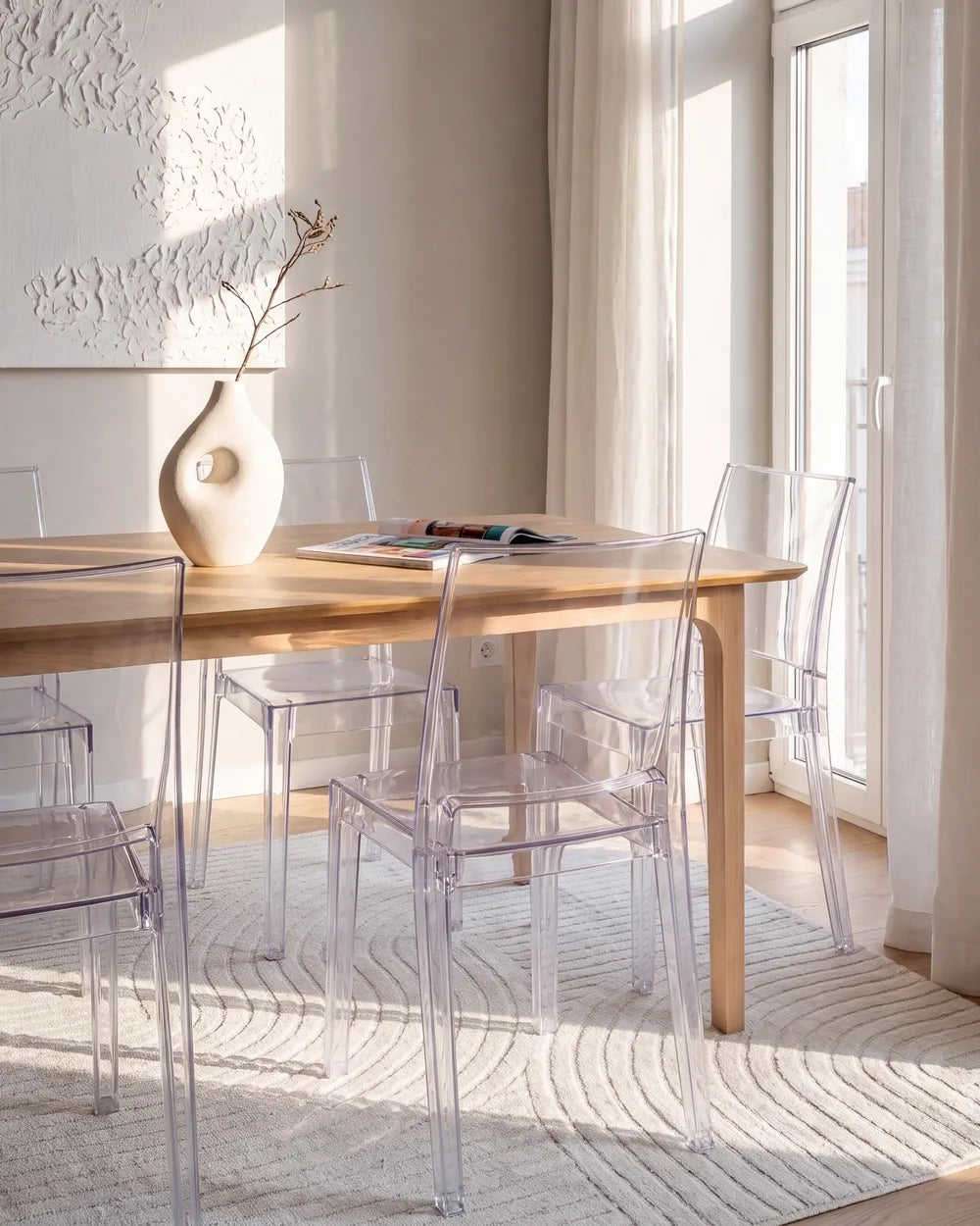A modern dining room scene with a light wood rectangular table surrounded by clear acrylic chairs, set on a textured cream rug near large windows with soft natural light.