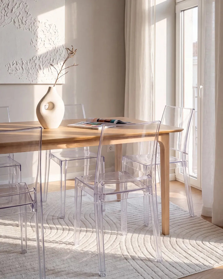 A modern dining room scene with a light wood rectangular table surrounded by clear acrylic chairs, set on a textured cream rug near large windows with soft natural light.