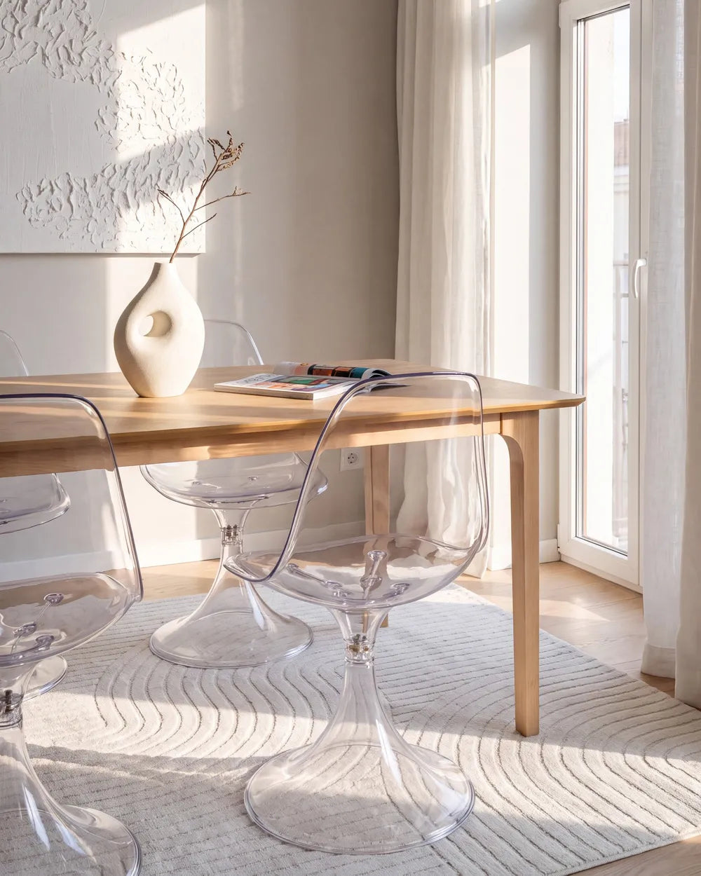 A modern dining setup featuring a light wood table and clear acrylic tulip-style chairs in a bright, sunlit room with a textured cream rug and a white ceramic vase on the table.