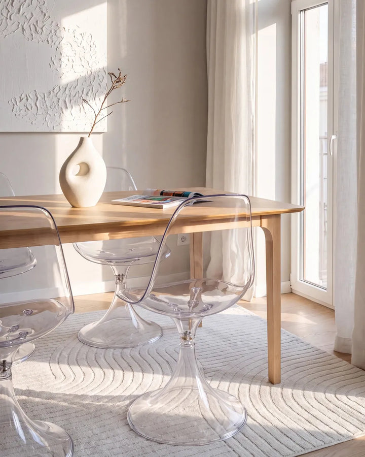 A modern dining setup featuring a light wood table and clear acrylic tulip-style chairs in a bright, sunlit room with a textured cream rug and a white ceramic vase on the table.