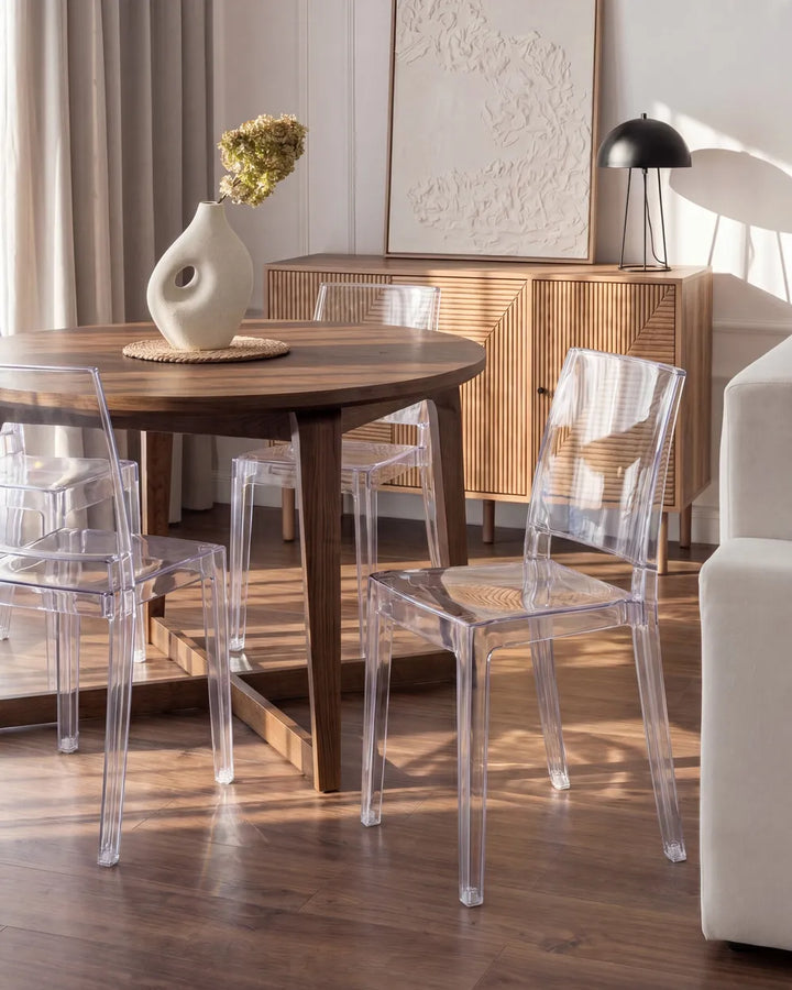 A round wooden dining table with four clear acrylic chairs in a modern dining room setting with warm wood floors and a wooden credenza.
