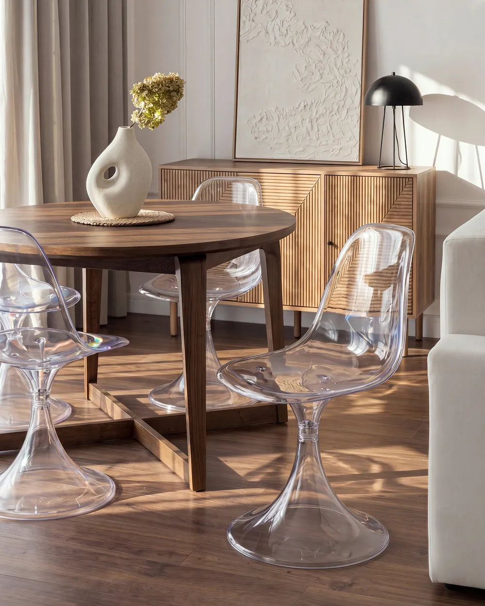 A modern dining room scene featuring clear acrylic tulip-style chairs around a round wooden table, with a wooden sideboard, vase, lamp, and framed wall art in the background.