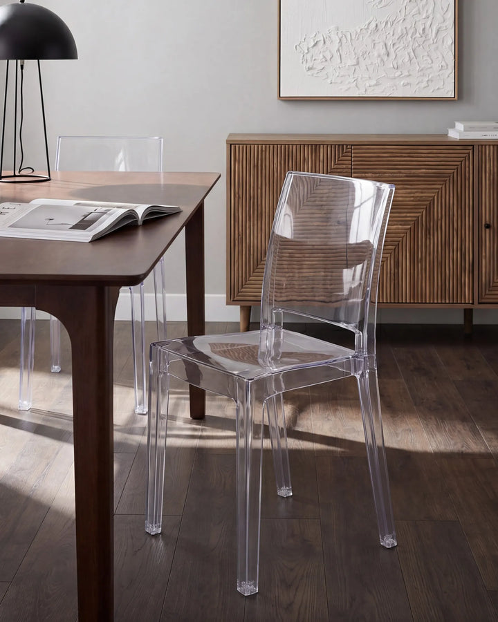 A clear acrylic dining chair in a modern dining room setting next to a dark wood table, with a wooden sideboard and dark wood floor in the background.