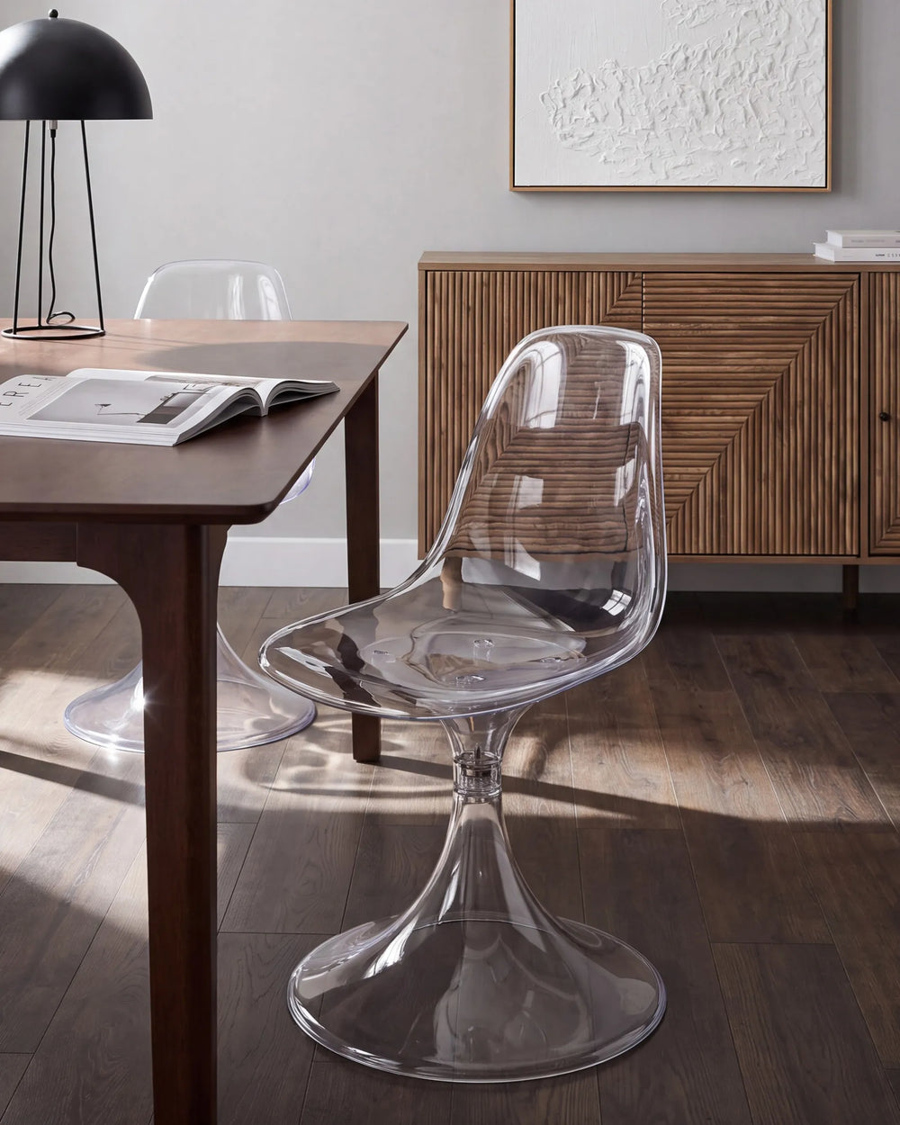 A clear molded plastic dining chair with a wide pedestal base, shown in a modern dining room setting next to a wooden table.