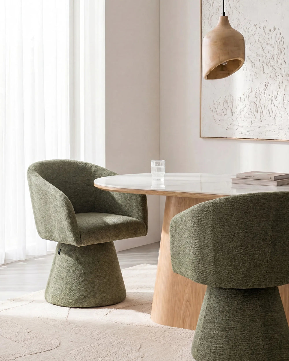 Two upholstered accent chairs in a muted sage green fabric arranged around a round light wood dining table in a bright, modern room.
