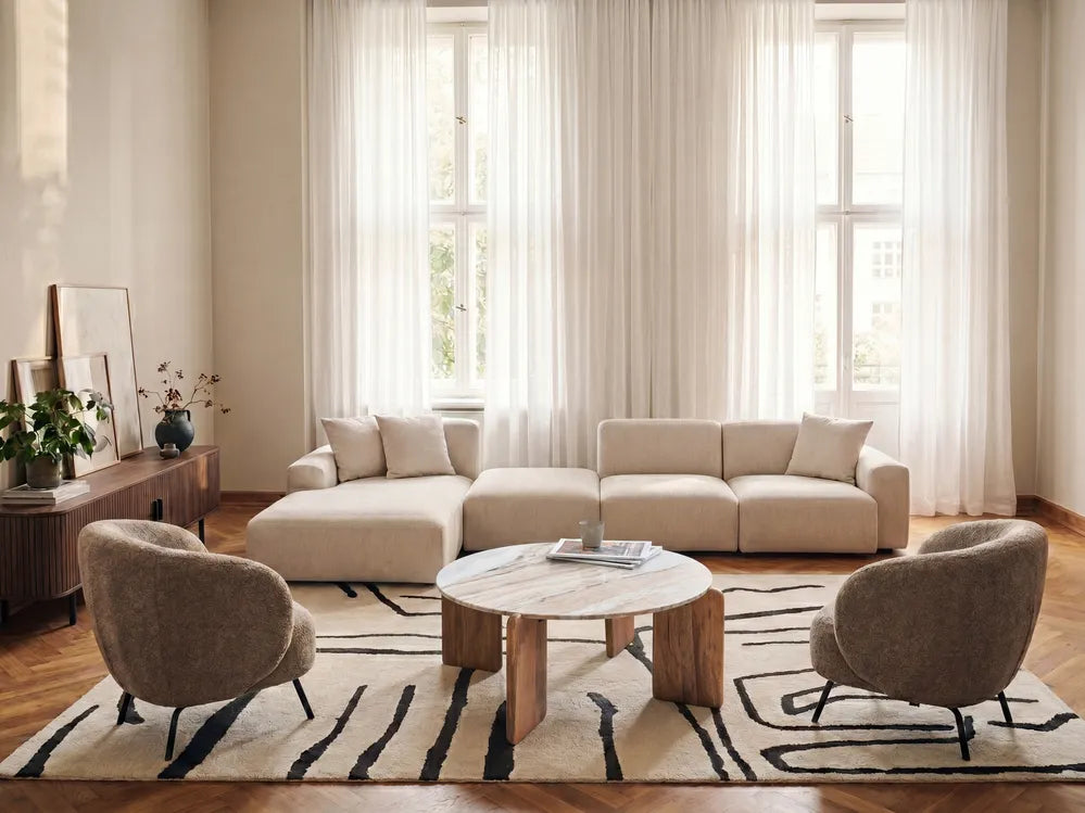A modern living room featuring a beige sectional sofa, two gray armchairs, a round marble-top coffee table, and a black-and-beige patterned rug in front of large windows with sheer curtains.