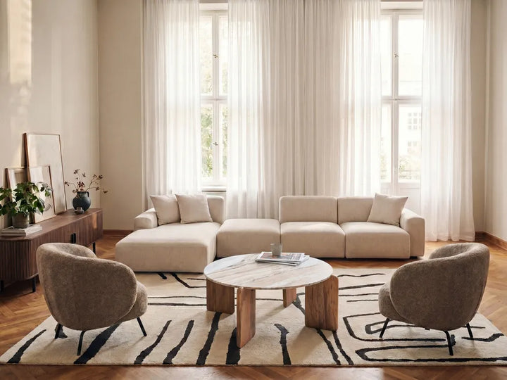 A modern living room featuring a beige sectional sofa, two gray armchairs, a round marble-top coffee table, and a black-and-beige patterned rug in front of large windows with sheer curtains.