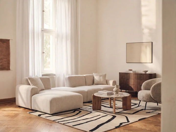 A modern living room featuring a cream-colored modular sectional sofa, a matching armchair, a low wooden coffee table, and a black and beige abstract patterned rug on a light wood floor.