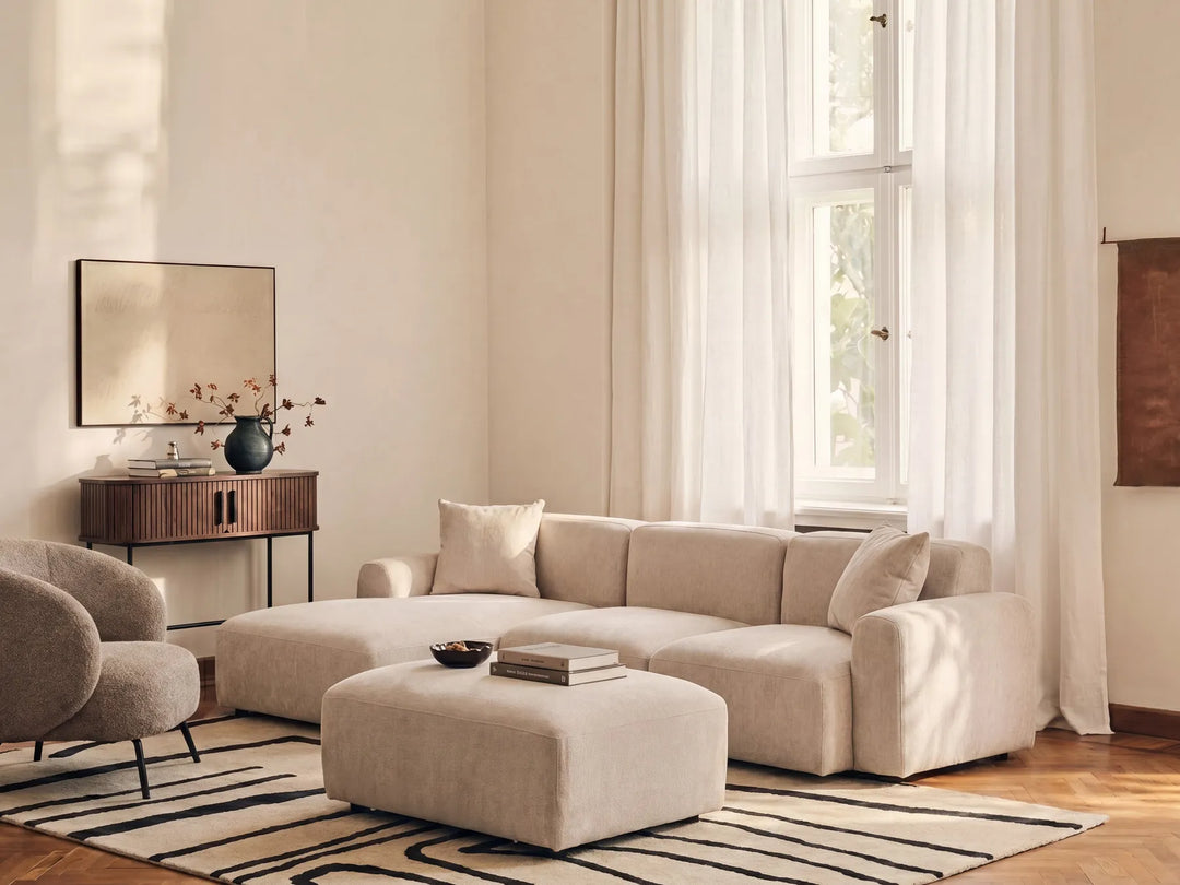 A modern beige sectional sofa with matching ottoman in a bright, minimalist living room with sheer white curtains, a striped rug, and a wooden sideboard.