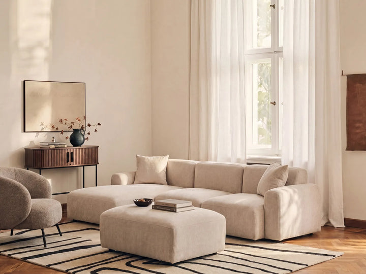 A modern beige sectional sofa with matching ottoman in a bright, minimalist living room with sheer white curtains, a striped rug, and a wooden sideboard.