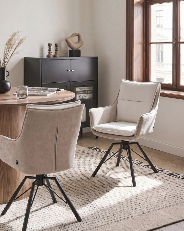 Two modern swivel dining chairs upholstered in light beige fabric with black metal legs, shown in a bright dining room setting next to a wooden table and window.