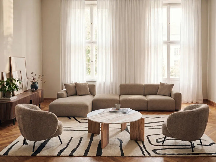 A modern living room setup featuring a beige sectional sofa, two matching armchairs, a round marble-top coffee table, and a black-and-beige patterned rug, set against large windows with sheer white curtains.