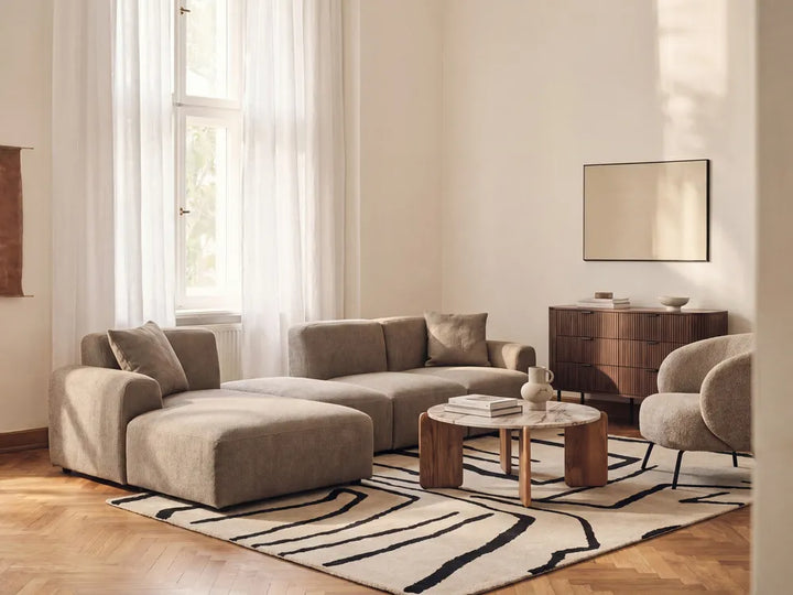 A modern living room featuring a beige sectional sofa with a chaise lounge, a matching armchair, a round wooden coffee table, and a black and beige abstract patterned rug on a light wood floor.