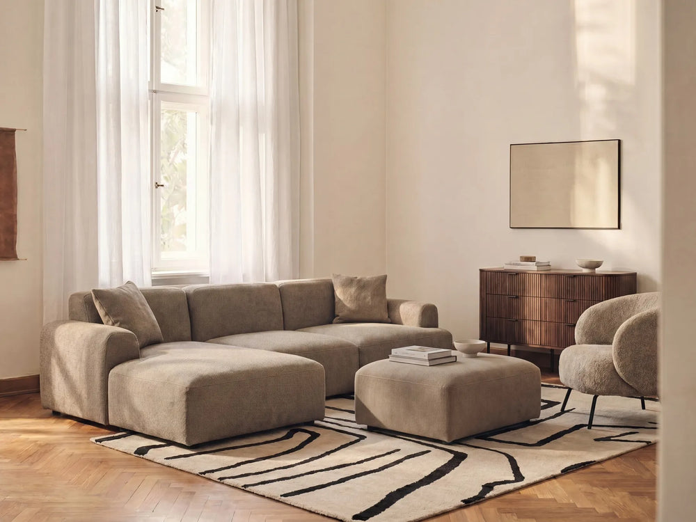 A modern beige sectional sofa with a chaise lounge, styled in a bright living room with a matching ottoman, patterned rug, and wooden sideboard.