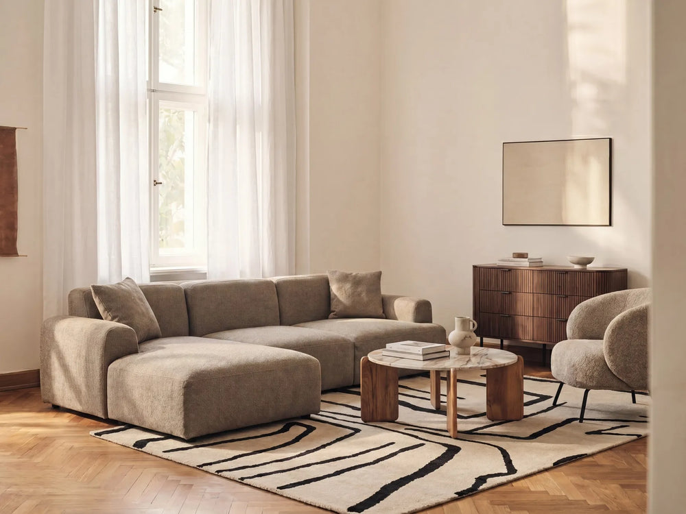 A modern living room featuring a beige sectional sofa with a chaise lounge, styled with a black and beige patterned rug, a round wooden coffee table, and a matching armchair.