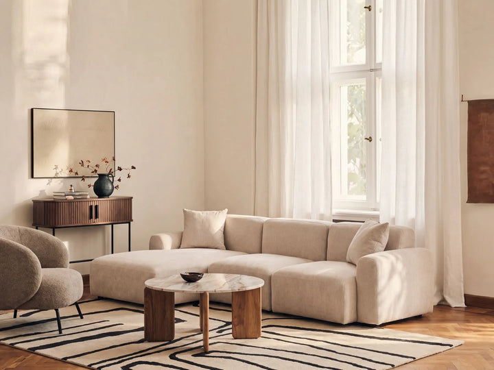 A modern beige sectional sofa in a bright, sunlit living room with white curtains, a striped rug, and a round wooden coffee table.