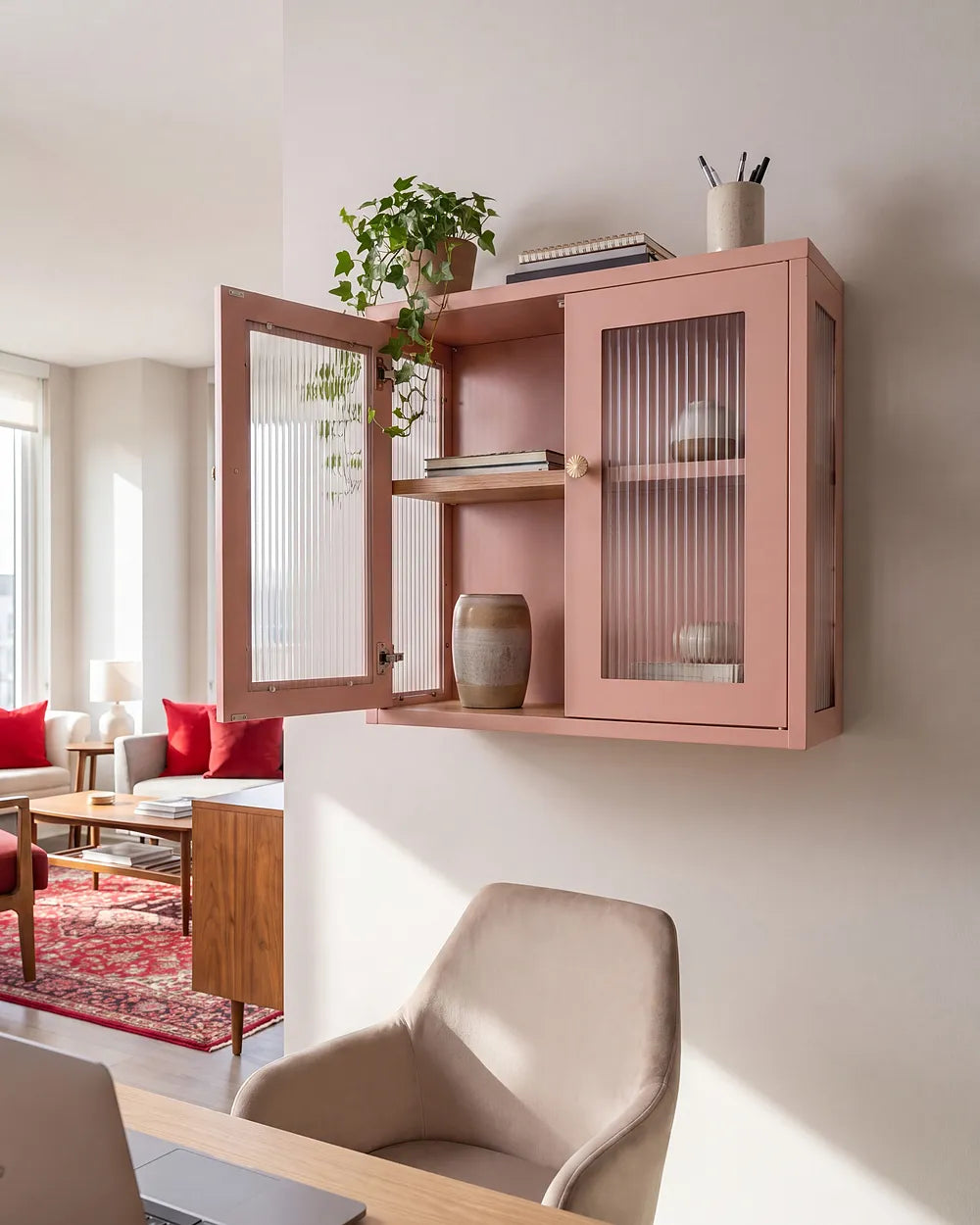 A pink floating wall cabinet with two glass-front doors and open shelving, mounted on a white wall in a modern living space. The cabinet is shown open with books, a vase, and a potted plant inside, with a chair and desk in the foreground.