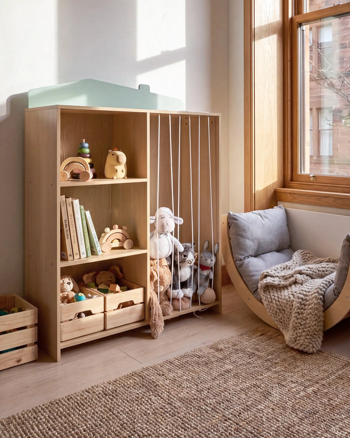 A light natural wood children's room furniture piece in a sunlit nursery, featuring open shelves with toys, a slatted side panel, and a mint green top trim, next to a wooden crescent chair with gray cushions and a knitted throw blanket.