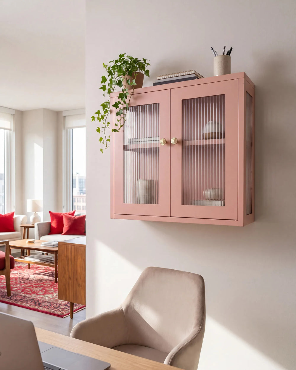 A pink wall-mounted cabinet with two ribbed glass doors and gold knobs, shown in a modern living space with a plant, chair, and window view.