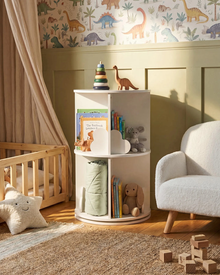 A white round corner bookshelf styled in a cozy children's nursery, holding books, a stuffed bunny, a plush elephant, and a small dinosaur toy. The nursery features dinosaur-patterned wallpaper, a wooden crib, a cream armchair, and a warm wooden floor.
