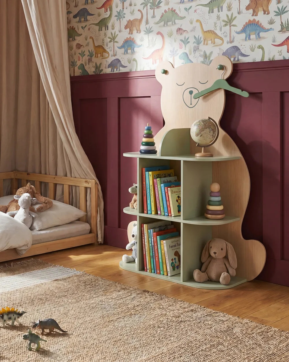 A children's bedroom scene featuring a light wood bear-shaped bookshelf with three open shelves, styled with books, toys, and a globe. The room has a beige canopy bed, a patterned dinosaur wallpaper, and dark burgundy wainscoting on the wall.