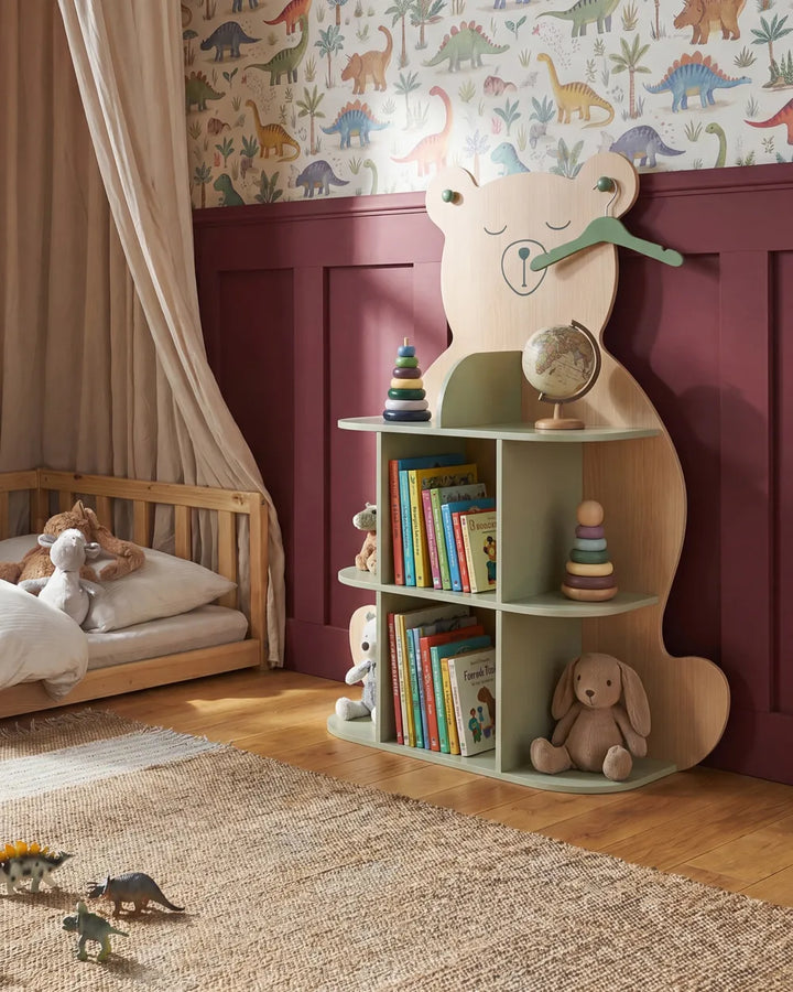 A children's bedroom scene featuring a light wood bear-shaped bookshelf with three open shelves, styled with books, toys, and a globe. The room has a beige canopy bed, a patterned dinosaur wallpaper, and dark burgundy wainscoting on the wall.