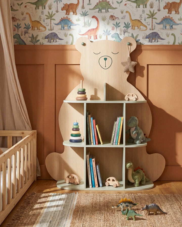 A wooden children's bookshelf shaped like a bear, styled in a nursery with dinosaur wallpaper, a crib, and toys on a rug.