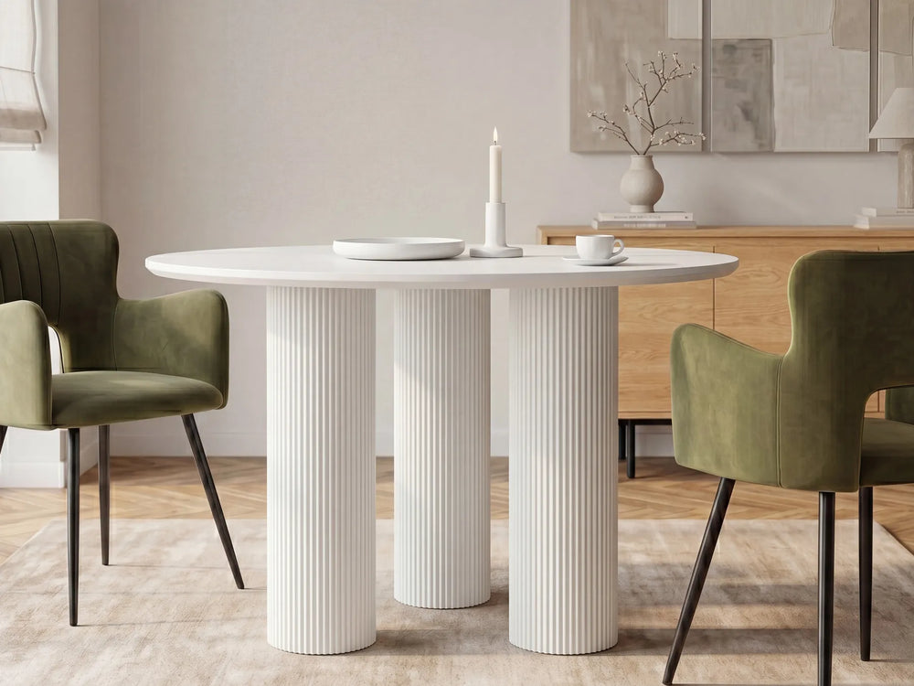 A modern round dining table with three fluted pedestal legs in white, styled in a dining room with two olive green velvet chairs, a wooden credenza, and neutral wall art.