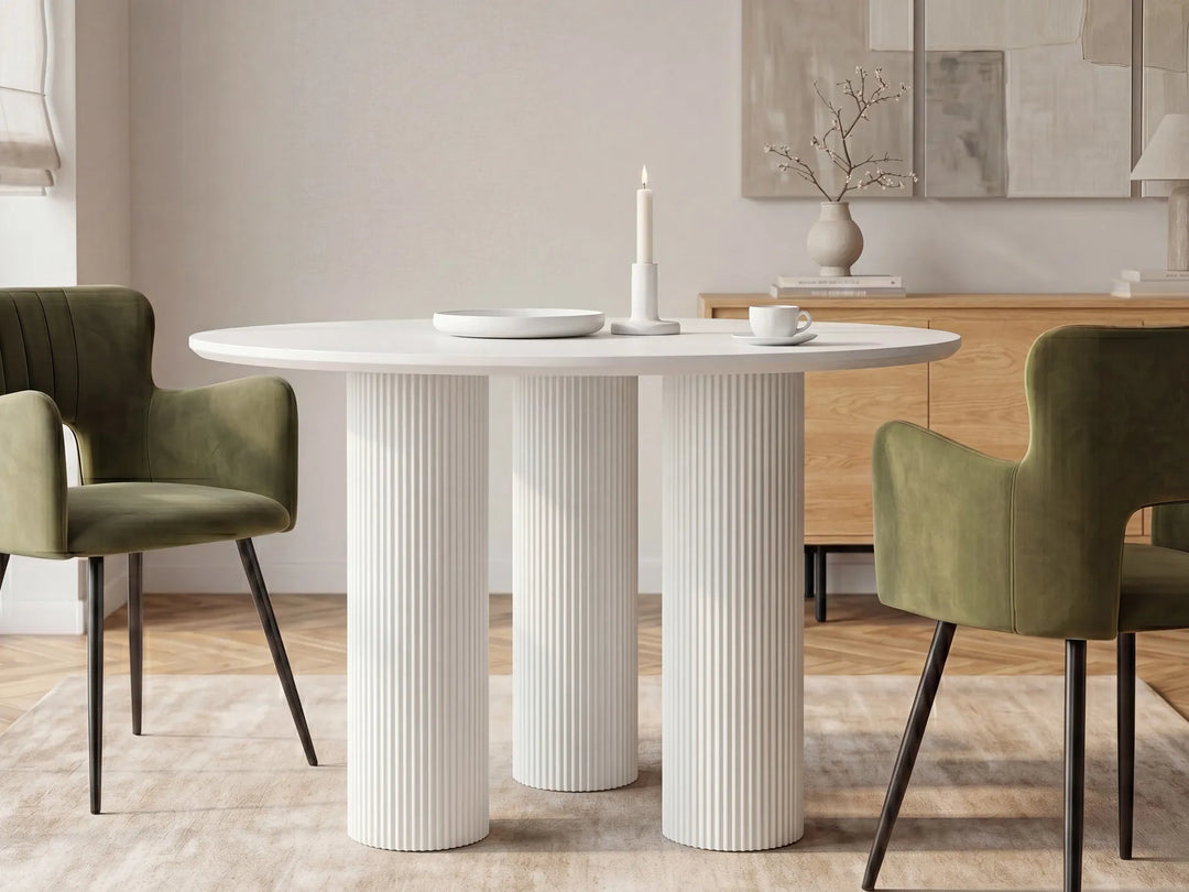 A modern round dining table with three fluted pedestal legs in white, styled in a dining room with two olive green velvet chairs, a wooden credenza, and neutral wall art.