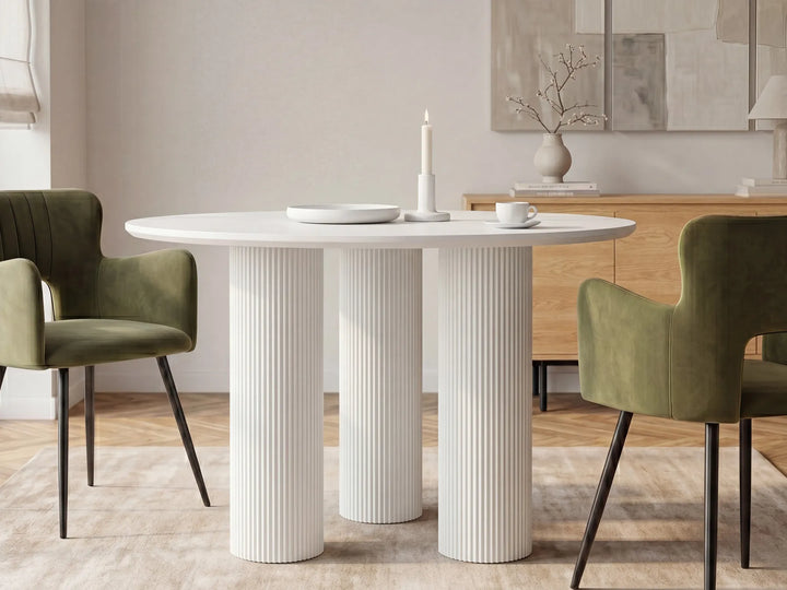 A modern round dining table with three fluted pedestal legs in white, styled in a dining room with two olive green velvet chairs, a wooden credenza, and neutral wall art.