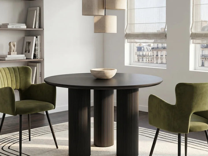 A modern round dining table with a dark wood top and fluted pedestal base, styled in a bright dining room with two green velvet chairs and a bowl on top.