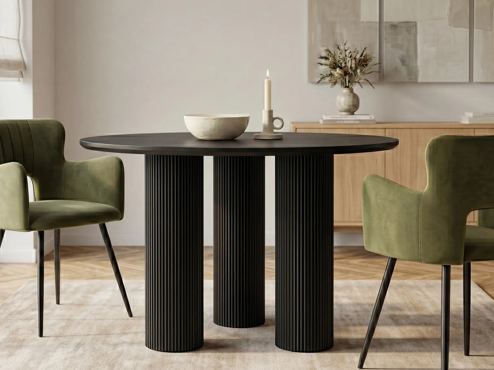A round dining table with three fluted pedestal legs in black, styled in a dining room with two green velvet chairs, a ceramic bowl, and a candle.