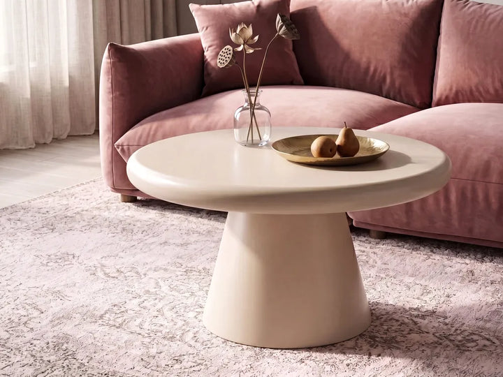 A round off-white coffee table with a tapered pedestal base, styled in a living room setting with a pink velvet sofa, a patterned rug, and a glass vase on top.
