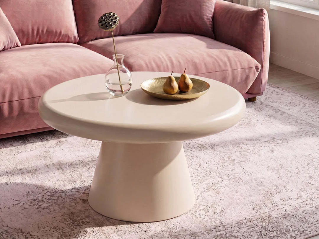 A round, low-profile coffee table with a smooth, glossy cream finish, styled in a living room setting with a pink sofa, a light rug, and decorative items on top.
