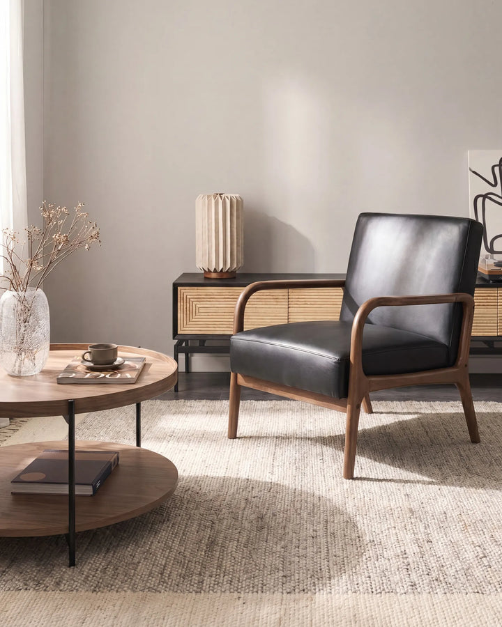A mid-century modern accent chair with black leather upholstery and a wooden frame, styled in a minimalist living room next to a round wooden coffee table.