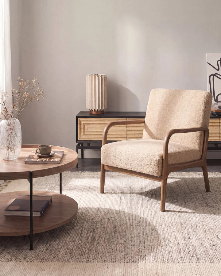A mid-century modern accent chair with textured beige upholstery and a natural wood frame, positioned in a styled living room next to a round wooden coffee table.