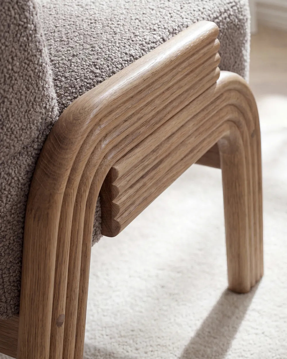 Close-up of a modern upholstered chair corner showing a curved light oak wood frame and textured taupe fabric seat on a light carpet.